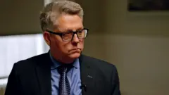 Kevin Hyland sentado, com terno escuro e óculos, durante entrevista com a BBC.