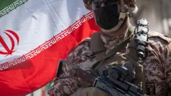 Membro armado da Guarda Revolucionária Islâmica do Irã com um fuzil AK-47 contra uma bandeira iraniana