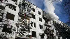 Bombeiro de pé sobre uma escada, ao lado de um edifício cinza de vários andares, com fumaça branca saindo dele