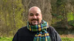 Daniel Marques de Araújo sorri para a câmera em meio a uma paisagem natural, com árvores e um lago ao fundo. Vestindo roupas escuras e um cachecol xadrez em tons de verde, azul e amarelo, ela aparece em primeiro plano, destacada pela luz suave do ambiente e pelo cenário tranquilo ao redor.