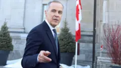 Марк Карни указывает пальцем в сторону объектива перед серым зданием с флагштоком, на котором флаг Канады