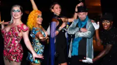 Laliberté aparece com cartola preta, casaco brilhante listrado em azul e preto e braços cruzados. Usa barba curta. À sua direita, duas mulheres com vestidos curtos e coloridos e meias arrastão pretas posam com os braços estendidos. Ao lado dele, uma mulher com vestido preto elegante segura uma taça de vinho e estende o braço esquerdo atrás dele. À sua esquerda, outra mulher, com vestido preto cintilante e luvas brancas com os dedos à mostra, faz um gesto em sua direção