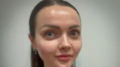 ਐਬੀ ਦੇ ਵਾਲ ਲੰਬੇ ਭੂਰੇ ਹਨ ਅਤੇ ਉਹ ਸਿੱਧਾ ਕੈਮਰੇ ਵੱਲ ਦੇਖ ਰਹੀ ਹੈ।