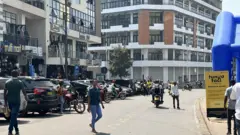 Abantu benshi bari mu muhanda wa kaburimbo urimo za moto n'imodoka nyinshi, zimwe ziparitse, iruhande hari inyubako ebyiri ndende.