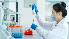 Un entorno de laboratorio donde una persona con bata blanca trabaja en una mesa de laboratorio. La persona lleva guantes protectores y mascarilla mientras utiliza cuidadosamente una pipeta para transferir líquido a un pequeño tubo de ensayo. En primer plano, sobre la superficie de trabajo, hay varias gradillas con tubos de ensayo que contienen líquidos de colores, dispuestas de forma ordenada. Al fondo se aprecian equipos científicos y estanterías, lo que sugiere un espacio de trabajo limpio y organizado.