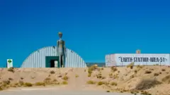 Araştırma merkezinin önünde dev bir gir uzaylı heykeli var. Yanındaki bir konteyner üzerinde büyük harflerle "Dünya-istasyon-51. Bölge" yazıyor.