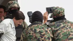 Em 22 de fevereiro de 2014, 'El Chapo' caminha com a cabeça baixa e as mãos para trás, cercado por forças de segurança em uniforme camuflado verde. Ele veste uma camisa branca com gola, tem cabelos pretos, com algumas entradas, e um bigode preto.