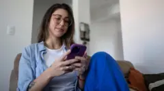 mulher jovem, de pele clara, cabelos escuros e óculos de grau no rosto, usando celular enquanto está sentada em um sofá