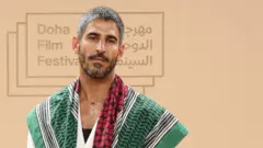 Una foto del periodista kuwaití‑estadounidense Ahmed Shihab‑Eldin, con el logotipo del Festival de Cine de Doha 2025 visible en el fondo. Tiene el cabello gris y una barba negra y gris, y viste una prenda con un patrón verde, rojo y blanco, además de un collar.