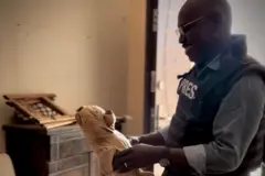 مراسل بي بي سي محمد عثمان وهو يقف داخل منزله الذي اضطر إلى مغادرته ويظهر في الصورة وهو يبتسم أثناء حمله لدُمية دب محشوة قديمة.