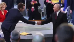 Pavel Zarubin y Vladimir Putin se dan la mano sobre una mesa en la que hay algunos papeles. Una mujer con un vestido rojo está sentada al fondo. Hay otras personas de pie detrás y delante de los dos hombres. Ambos llevan trajes oscuros y Putin lleva una corbata burdeos.