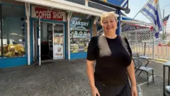 Martha Machado posa ante la cámara delante de su panadería uruguaya en Elizabeth, Nueva Jersey. Viste una camiseta negra manga corta y un pantalón negro, y tiene los labios pintados de rojo. A su izquierda, detrás, se pueden ver las banderas de Uruguay y de Estados Unidos.