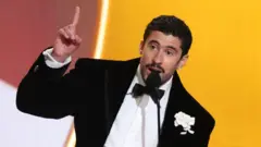 ひげを蓄えた男性が、マイクの前で話している。タキシードにボウタイ、胸ポケットに白い花を着けている。右手の人差し指を挙げている