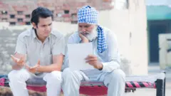ਇੱਕ ਨੌਜਵਾਨ ਅਤੇ ਬਜ਼ੁਰਗ ਮੰਜੇ ਉੱਪਰ ਬੈਠੇ ਕੋਈ ਕਾਗਜ਼ ਦੇਖ ਕੇ ਪਰੇਸ਼ਾਨੀਵ ਦੀ ਮੁਦਰਾ ਬਣਾਉਂਦੇ ਹੋਏ