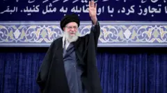 Хаменеї в традиційному чорному та темно-синьому одязі, яка стоїть на сцені та піднімає руку в жесті вітання. За нею – великий синій банер із декоративним орнаментом та арабським написом. Сцена виглядає як офіційний або релігійний виступ у залі з оформленою трибуною.