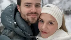 Two people standing close together in the foreground, wearing padded winter jackets, with one in a dark green coat and the other in a white coat and knitted headband. Snow is visible on the clothing. Behind them are snow-covered trees and branches.