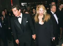 John F. Kennedy y Carolyn Bessette. Él viste esmoquin negro y ella un traje negro. Caminan tomados de la mano, mientras detrás de ellos se ven otras personas no identificadas vestidas de etiqueta.