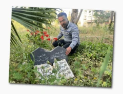 ডোরাকাটা ধূসর কলারওয়ালা পোশাক পরা মোহাম্মদ আরবি লেখা সম্বলিত সাদা-কালো কবরের পাশে হাঁটু গেড়ে বসে আছেন। পাশে লাল ফুল লাগানো রয়েছে