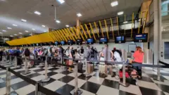 Passageiros em fila nos balcões de check-in do Aeroporto de Congonhas, com malas e carrinhos de bagagem, diante de painéis de atendimento de companhias aéreas.