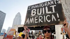 Una manifestante con lentes oscuros y una pancarta que dice "Immigrants built America!"