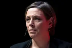 Retrato de Jess Phillips em um palco ou evento com fundo escuro. Ela veste um casaco escuro e usa vários brincos