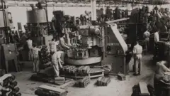 A imagem em preto e branco mostra o interior de uma fábrica com diversos operários trabalhando ao redor de grandes máquinas industriais. Os trabalhadores usam macacões claros e manipulam peças metálicas espalhadas pelo chão e sobre bancadas, enquanto equipamentos de grande porte ocupam quase todo o espaço do galpão.