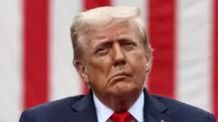 Ifoto ya Perezida w'Amerika Donald Trump igaragaza intugu n'umutwe. Arareba muri 'camera', yambaye ikositimu y'ubururu, ishati y'umweru na karuvati itukura. Akoze ikimenyetso n'umunwa. Inyuma ye hari igitambaro kitagaragara neza cy'amabara y'umutuku n'umweru. 