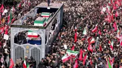 د زرګونه خلکو ترمنځ‌ یو لوی موټر روان برېښي چې تابوتونه پکې د ایران په بیرغ کې پوښل شوي ایښي دي. 