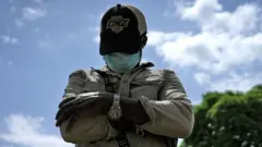 James (abụghị ezigbo aha ya) na-eleanya n'ala ka ọ looks down as he poses for a photo with his arms folded wearing a wrist watch, baseball cap, face mask, trousers and a khaki shirt. He is standing outside and a blue sky can be seen in the background