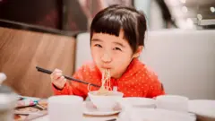 A girl in a red, polka-dotted shirt smiles for the camera while tucking into a bowl of noodles