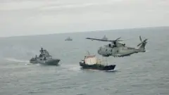 Над морем летить військовий вертоліт, нижче в воді рухаються кілька кораблів, серед них військові судна та одне цивільне. Море спокійне, небо хмарне; сцена виглядає як військове патрулювання або супровід у відкритому морі.