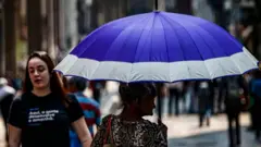Uma senhora se protege do sol com uma sombrinha roxa e branca.