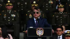 Gustavo Petro durante discurso. Ele usa terno e óculos escuros. Atrás dele há militares