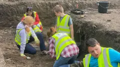 Estudiantes con chalecos reflectantes ayudan en la excavación al oeste de la iglesia de la Abadía de Margam, supervisados por adultos