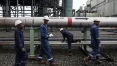 Gas workers dress for protective gear waka past large pipeline labelled 'natural gas'.