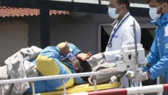 Dawa Sherpa on a stretcher acompanied by two doctors. He is wearing  a blue and yellow mountian jacket