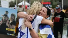 две женщины с израильскими флагами на плечах обнимают друг друга на фоне плакатов с фотографиями семей заложников и погибших