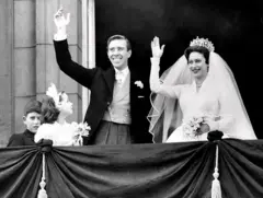 Una pareja de recién casados, él de sacoleva y ella de blanco con velo y una corono, saludan desde un balcón con los brazos en alto.