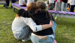 Two teenage girls sit with their arms around each other, hugging, while sitting in front of a camp fire