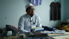 Soe Nay Oo mira con preocupación a lo lejos en su dormitorio, con una gorra y uniforme blancos de imán y gafas de montura negra. Al fondo se ven su cama y camisetas colgadas en la pared.