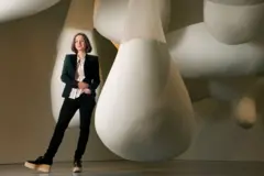 Sarah Thornton, vestida con una chaqueta y un pantalón negros y una blusa blanca, al lado de una obra de arte que consiste en formas gigantes de color blanco