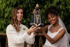 Fotografia colorida mostra duas noivas sorrindo e segurando juntas uma estátua de Nossa Senhora Aparecida. A mulher da esquerda, loira, usa um camisa branca com uma flor na lapela. A mulher da direita, negra, usa um vestido de noiva