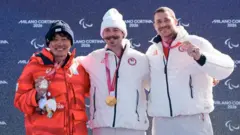 3人が横並びになっている。小栗は赤いウエアに黒っぽい帽子、他の2人はそろいの白いウエアで、エリオットは白い帽子をかぶっている。全員笑顔。小栗とシュルツは片手でメダルを持ち、胸の前で見せている