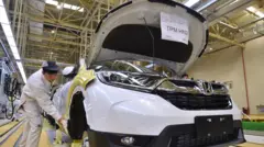 Un automóvil Honda blanco que se fabrica en una fábrica con un trabajador a la izquierda de la imagen con un uniforme blanco y una gorra.
