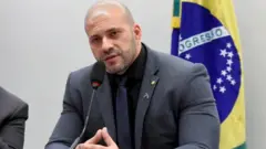 Daniel Silveira em sala da Câmara, falando em microfone e sentado, com bandeira do Brasil atrás