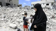 Yıkılan bir binanın önünde duran siyah çarşaflı bir kadın ve arkası dönük bir kız ve erkek çocuğu.
