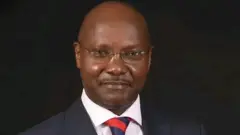 Man wey dey look di camera, e wear glass, white shirt, black suit and bald head