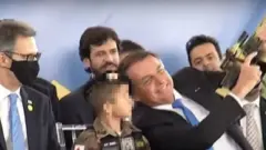 Reprodução de vídeo no Youtube mostra Bolsonaro sorrindo e apontando arma para cima, ao lado de criança e homens