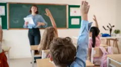 Bir sınıfta ilkokul öğrencisi bir erkek çocuk iki kız çocuk el kaldırıyor. Arkada bir kadın öğretmen elinde kağıtla konuşuyor. 