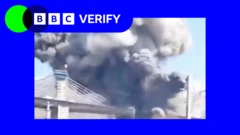 Smoke rises from a strike on the B1 Bridge in Iran. The photo is imposed over the BBC Verify colours and branding. 